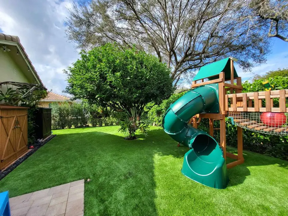 A backyard with Southwest Greens of Tucson's vibrant artificial grass features a wooden play structure with a green spiral slide. A large leafy tree stands nearby, providing shade. The sky is partly cloudy, with surrounding plants and a house visible, creating an inviting space for relaxation and fun.