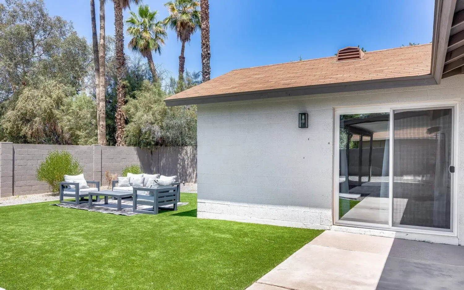 A backyard in Catalina Foothills with artificial grass installation showcases a white house with a sliding glass door. The modern outdoor seating features gray furniture and white cushions on a patterned rug, framed by tall palm trees and a block wall fence.