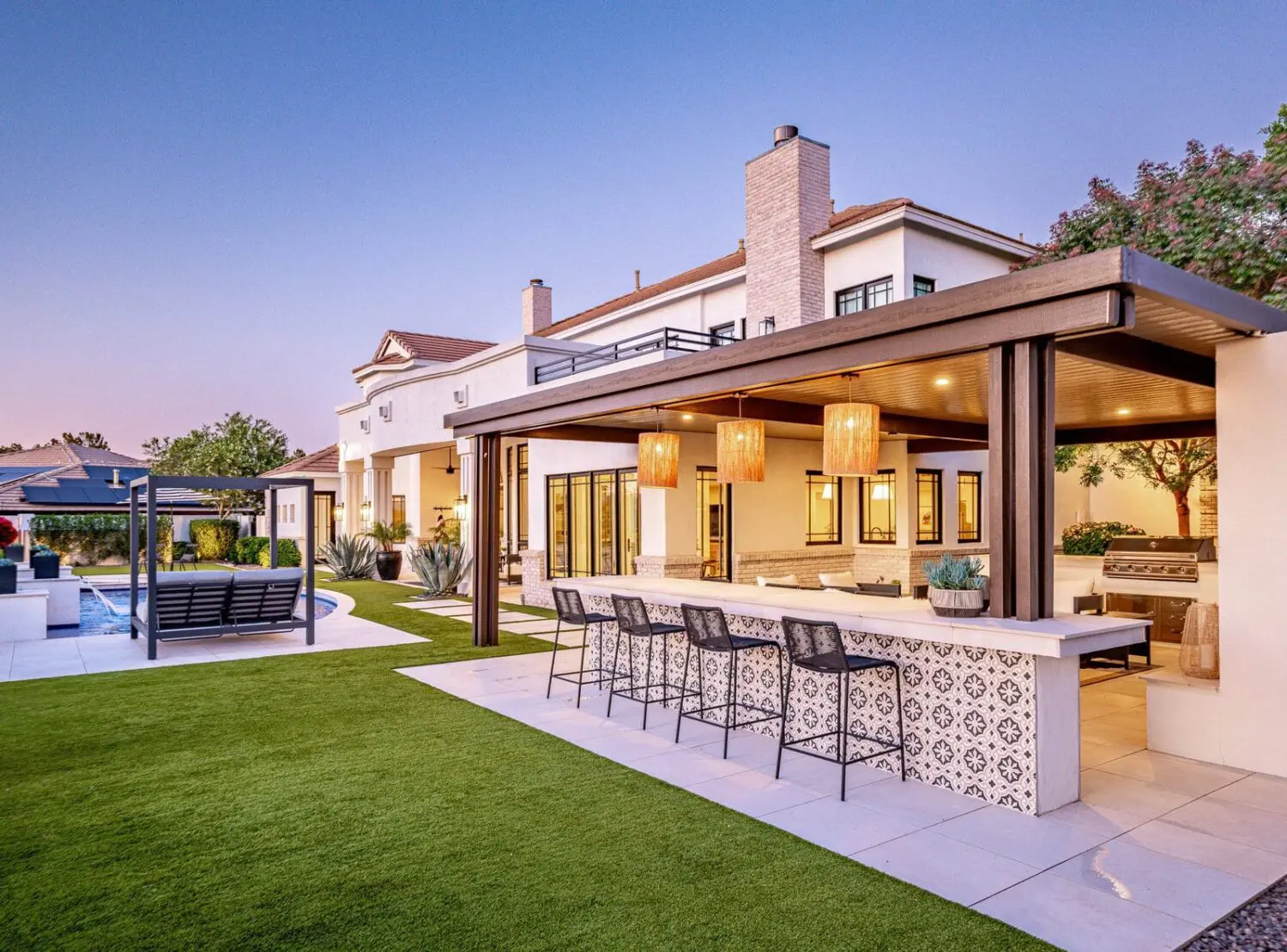 Luxury modern house in the Catalina Foothills boasts a spacious patio with a bar counter, patterned tiles, and overhead lights. A large swimming pool and lounge chairs rest atop well-manicured quality turf solutions. The clear sky enhances the serene ambiance.