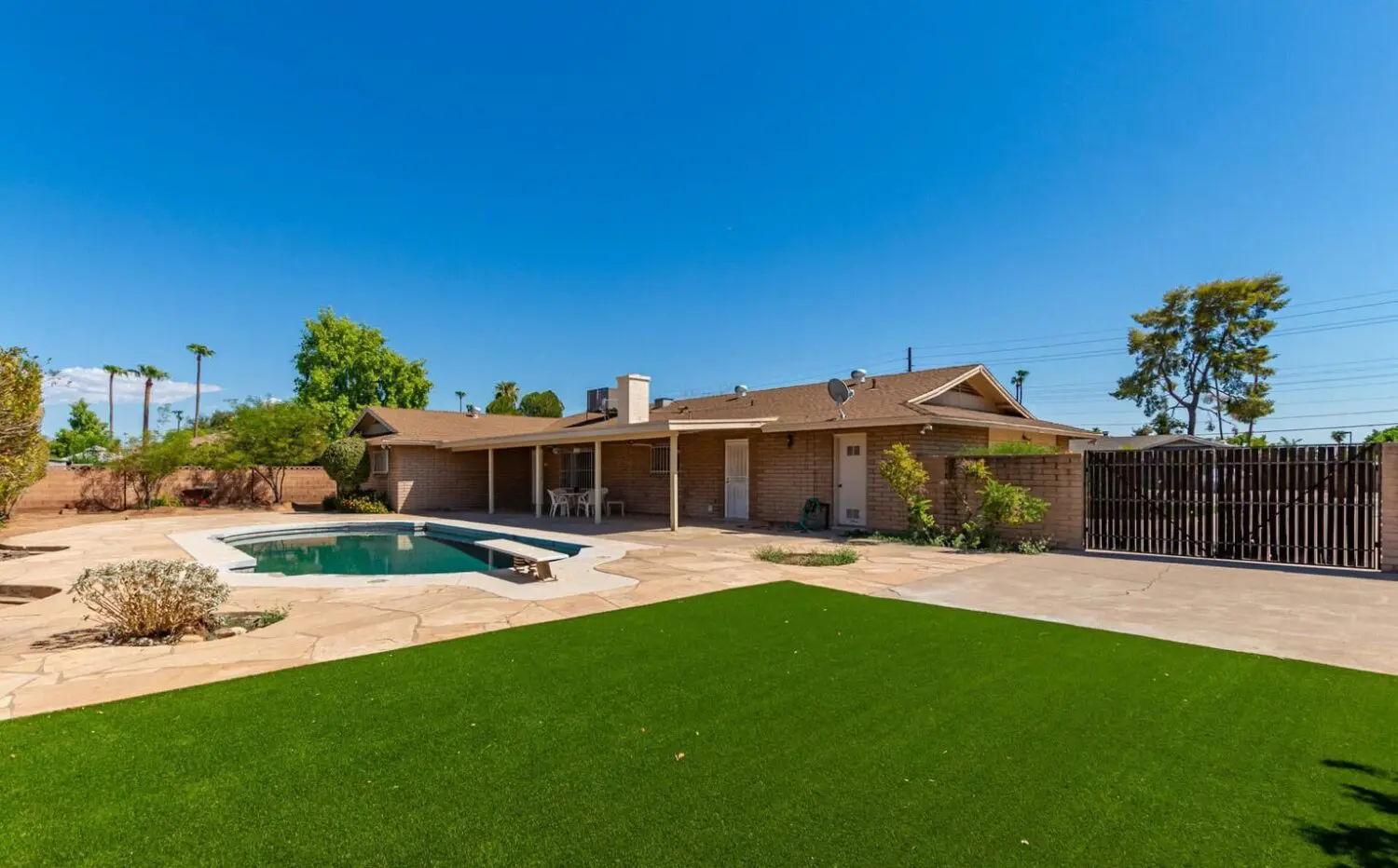 A backyard in Marana, AZ, featuring a single-story brick house with a chimney and covered patio. The area boasts a swimming pool surrounded by stone patio, vibrant artificial turf, and wooden fence under a clear blue sky.