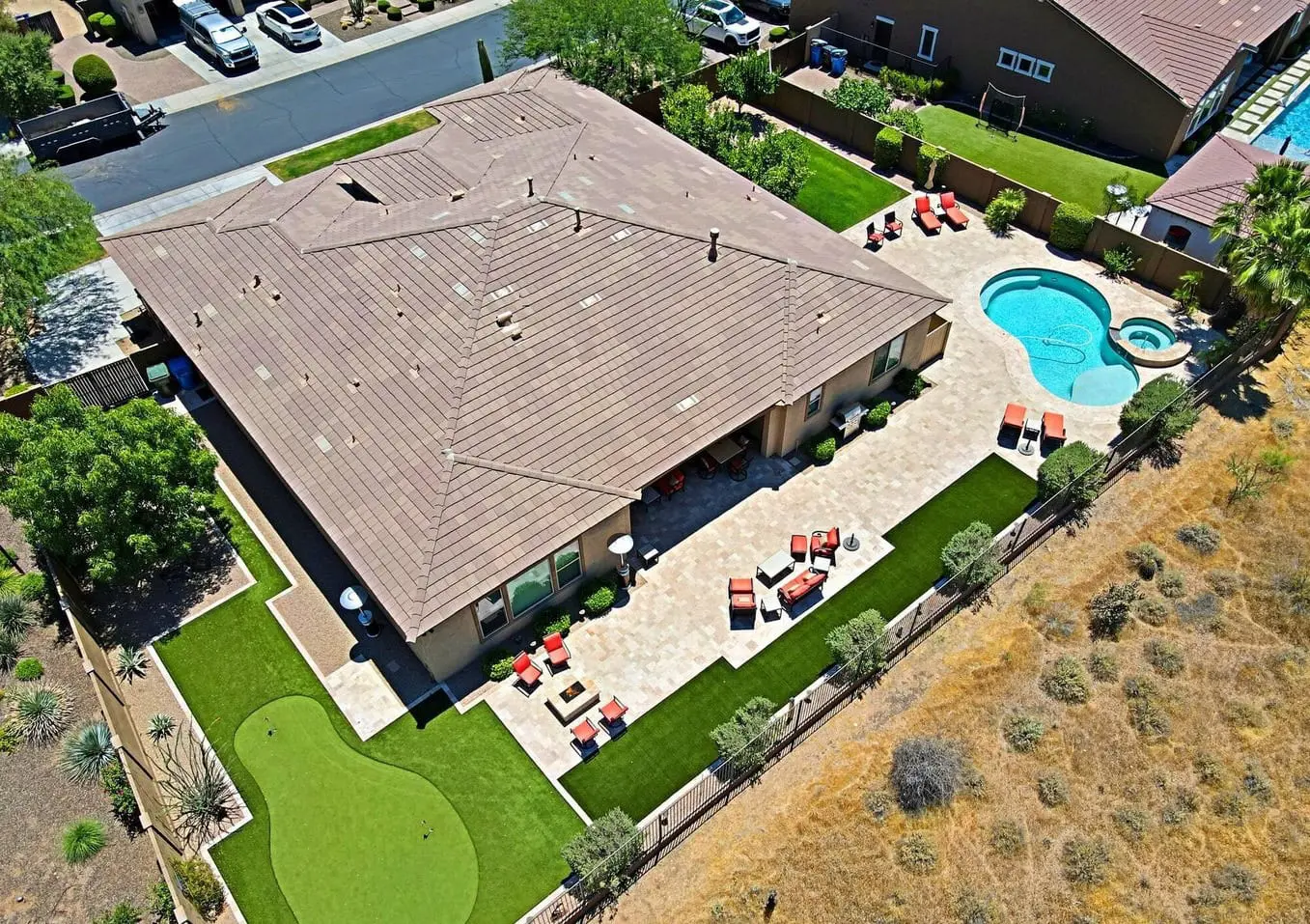 Aerial view of a large house with a tiled roof in Marana, AZ. The backyard boasts a patio with seating, swimming pool, hot tub, and a putting green set on lush artificial grass. Surrounded by trees and dry grasslands, neighboring houses and streets are visible in the distance.