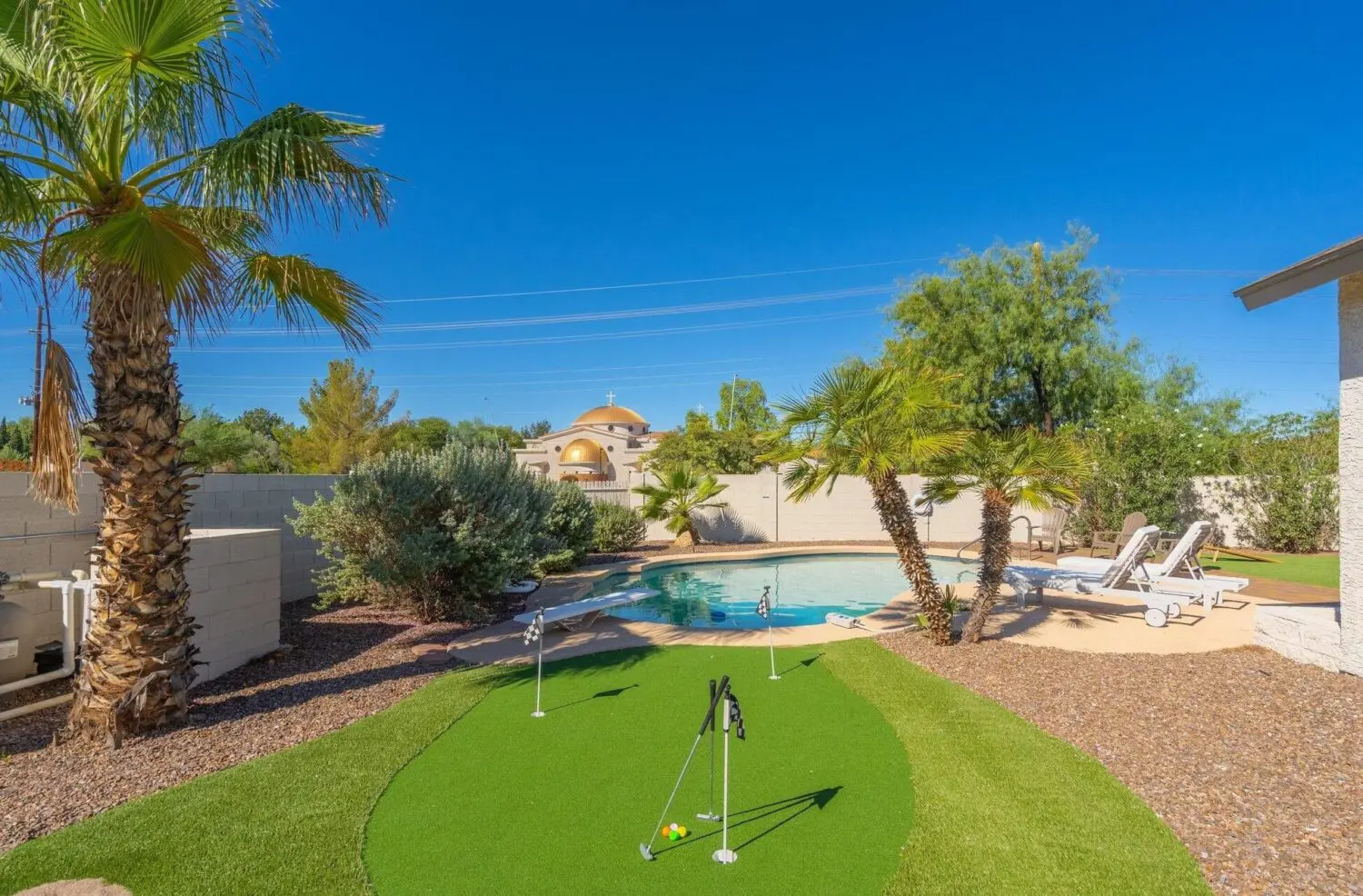 In Marana AZ, a backyard oasis boasts a small putting green of lush artificial turf with three flags, bordered by palm trees and shrubs. In the background, there's a shimmering swimming pool with lounge chairs basking under the clear blue sky.