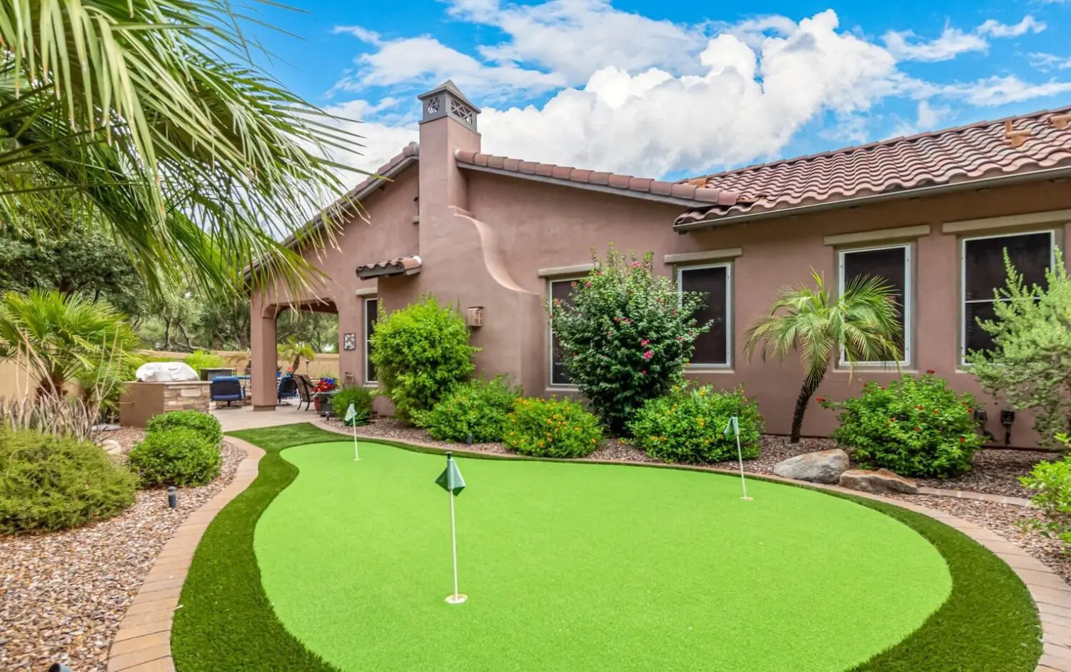 A backyard scene in the Catalina Foothills showcases a putting green with lush turf solutions, nestled among vibrant plants and shrubs. The house boasts a stucco exterior and tile roof under a blue sky dotted with clouds, while a grill peeks out from the left.