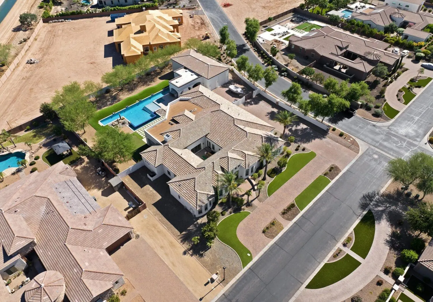 Aerial view of a suburban neighborhood in Catalina Foothills featuring a large house with a pool, landscaped yard, and expertly installed artificial grass. Surrounding homes boast spacious lots, greenery with turf solutions, and a mix of completed and under-construction buildings. Roads and driveways are visible.
